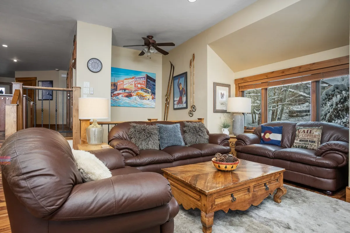 A spacious living room featuring brown leather sofas and a wooden coffee table, set against large windows that reveal a snowy downtown landscape. Vibrant artwork adds local character while natural light streams in, suggesting a cozy environment with a view to the outside world.