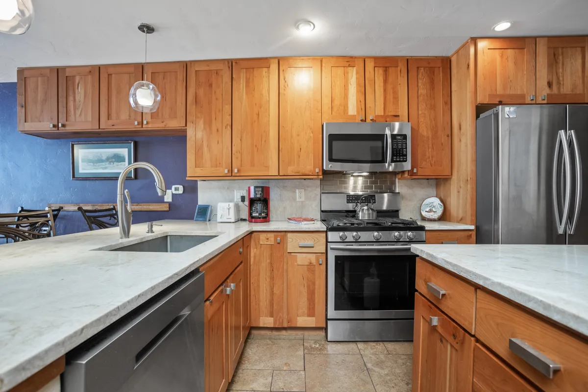This image features a bright kitchen with a large island, stainless steel appliances, and wooden cabinetry. Morning light spills across the countertops, creating a cozy and inviting atmosphere perfect for making coffee or preparing meals. The scene suggests comfort and daily living, with visible areas for both cooking and socializing.