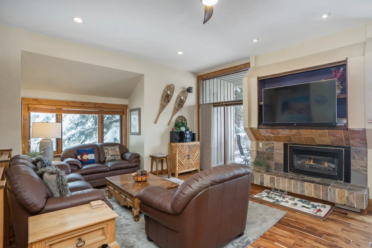 A warm, inviting living room with a fireplace as the focal point, flanked by large windows that offer views of the outdoors. Comfortable seating is arranged for group gatherings, and a wooden coffee table sits at the center, creating a cozy, communal atmosphere ideal for relaxing après-ski or after a day out.
