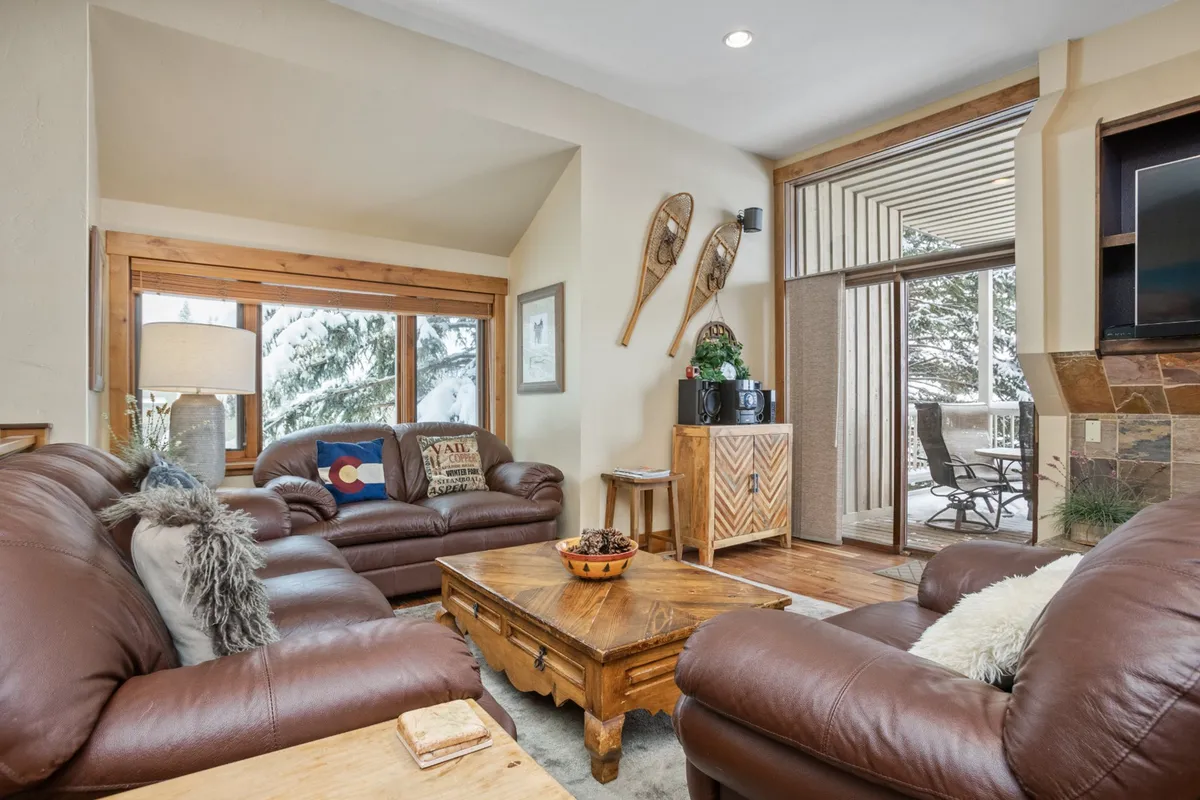 This image presents a welcoming living room with comfortable leather sofas arranged around a coffee table. Large windows bathe the space in natural light, and decorative snowshoes on the wall hint at the mountain setting. Lamps add ambient lighting, and the overall setup encourages relaxation, socializing, and enjoying views.