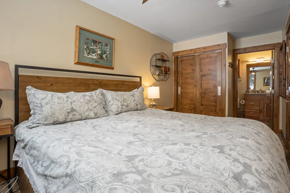 A spacious, beautifully lit master bedroom featuring a plush king-sized bed with decorative pillows, a nightstand with a lamp, wall artwork, and a wooden cabinet. The tranquil atmosphere and thoughtful styling suggest restful nights and a private retreat, perfectly aligning with the master suite description.