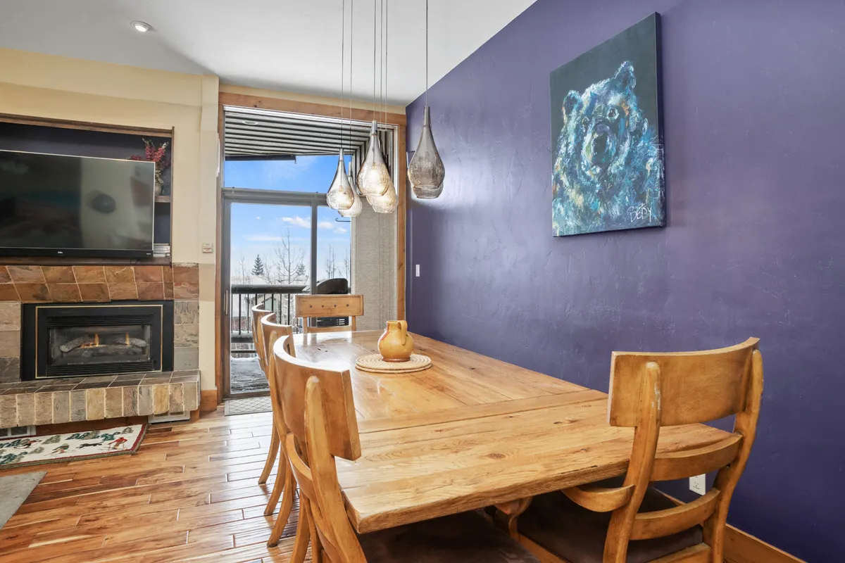 A modern dining area with a prominent dining table sits beneath stylish light fixtures, with a striking bear painting adding character. The fireplace glows in the background, and windows frame the scene, suggesting both warmth and scenic surroundings. The table is set for gatherings and meals.