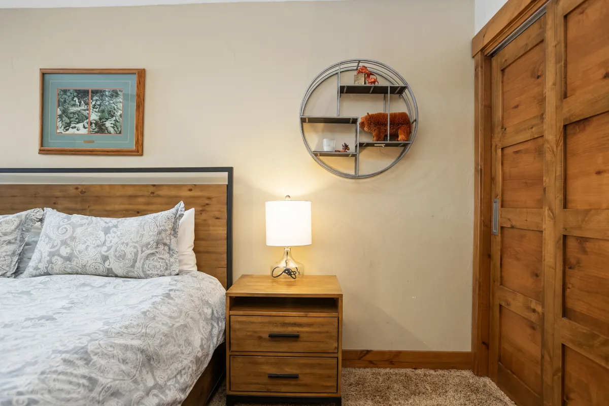 A cozy bedroom scene with a queen bed dressed in inviting bedding, a nightstand with a lamp, and a decorative shelf with curated items. The room is filled with natural light, emphasizing comfort and the welcoming atmosphere ideal for families or groups.