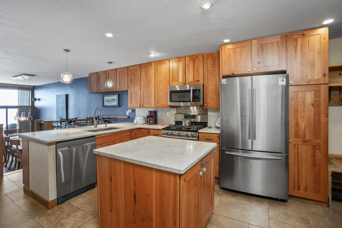 The kitchen shines with stainless steel appliances, a generous island, and warm wood cabinetry. The space is open and connected, suggesting effortless movement between cooking, gathering, and dining. Modern pendant lights and natural textures create a contemporary, welcoming vibe.