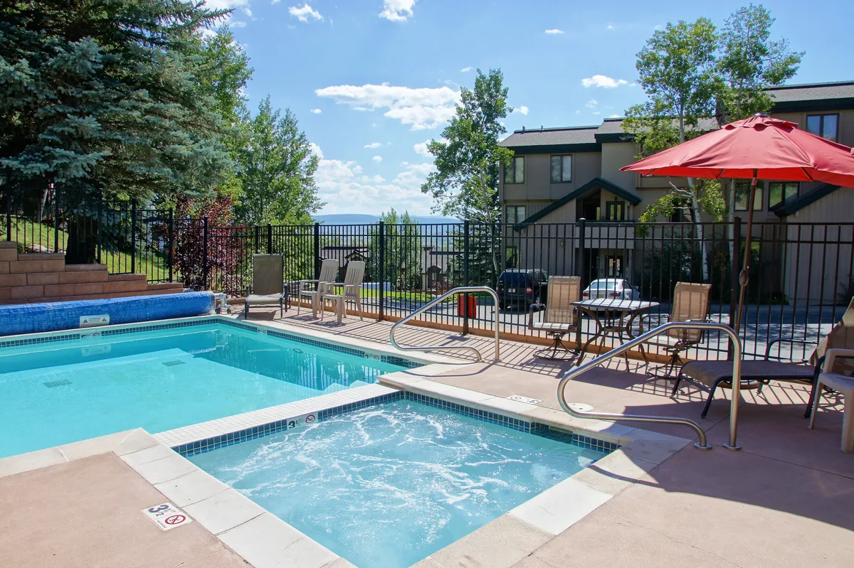 The image showcases the condo's inviting outdoor heated pool and bubbling hot tub surrounded by comfortable seating and umbrellas, perfect for relaxing after a day on the Steamboat Springs slopes. The scene is set for sunset swims, poolside barbecues, and stargazing under the Colorado sky, offering an atmosphere of warmth and relaxation.