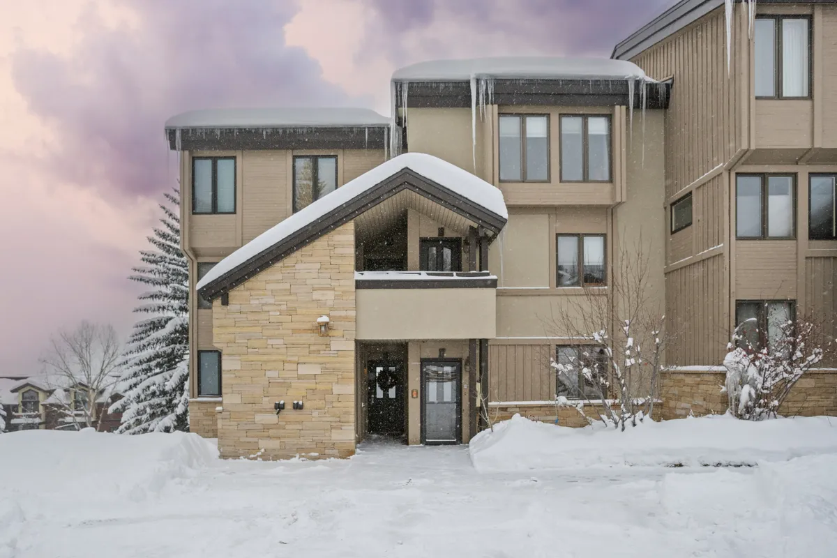 A snow-covered entrance to the building, featuring a stone facade, icicles hanging from the roof, and warmly lit windows that hint at the cozy interior. Snow blankets the ground, creating a classic winter lodge atmosphere close to the slopes.