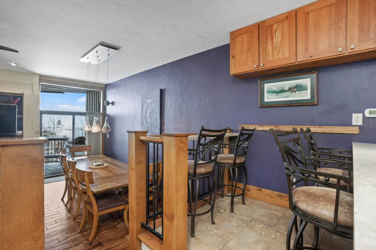 A bright interior dining area with a table and bar stools near large windows offers inviting views of the outdoors. The sunlight flooding the space and window vistas create a strong connection between indoor comfort and the appeal of the surrounding environment.
