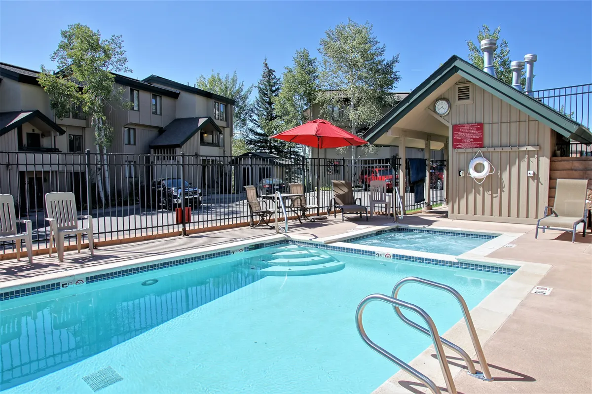 A vibrant outdoor scene featuring a sparkling swimming pool surrounded by red umbrellas and comfortable lounge chairs. Lush trees encircle the pool area, providing a peaceful, private setting for relaxation, swimming, and sunbathing.