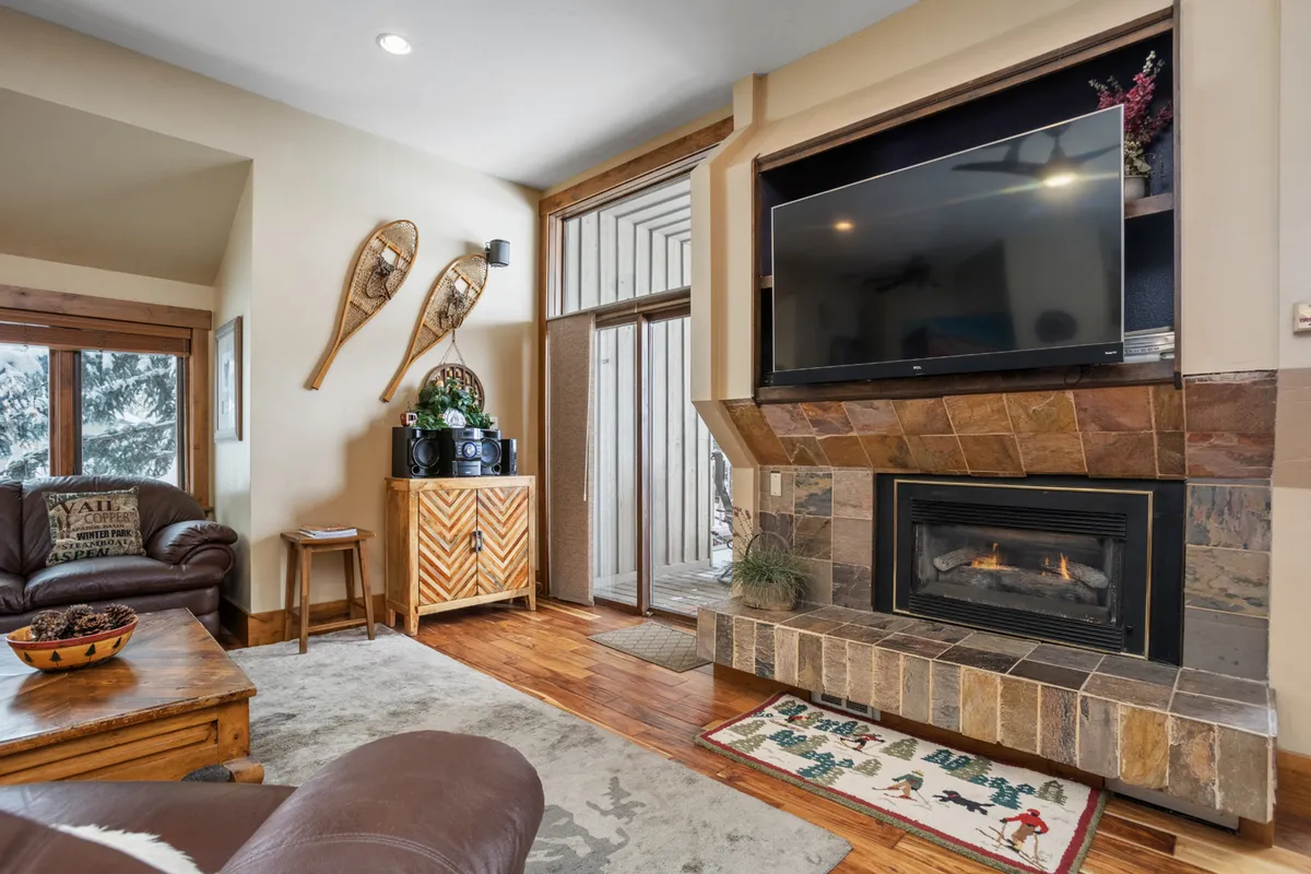 A living area centered around a fireplace, with wooden furniture, a television, and windows that let in natural light. The setting is designed for comfort and socializing, perfect for quiet evenings, movie nights, or inviting friends over. The visible windows give a sense of connection to the outdoors, tying the interior to the broader environment.