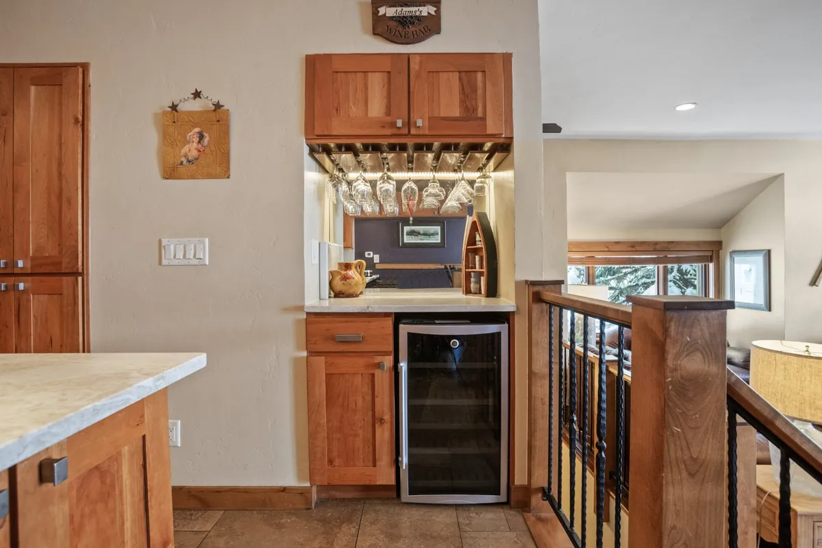 A stylish nook with a wine fridge, hanging wine glasses, and wood cabinetry set the stage for casual gatherings. The area exudes warmth and comfort, with an art piece adding a personal touch, suggesting a space to unwind with a beverage or entertain friends after a day of remote work or exploration.